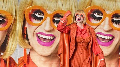Drag Queen Delta wearing a plaid orange dress suit and bright orange glasses