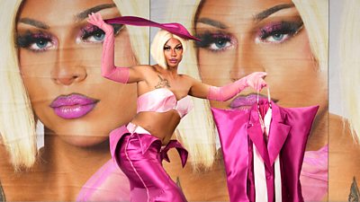 Drag queen Paris in a pink skirt, a pink top and pink fascinator, they are holding a bright pink suit bag