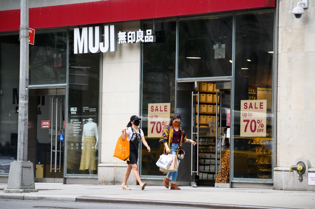 Two women wearing face masks and carrying shopping bags walk past a MUJI store with a "SALE up to 70% OFF" sign in its window.