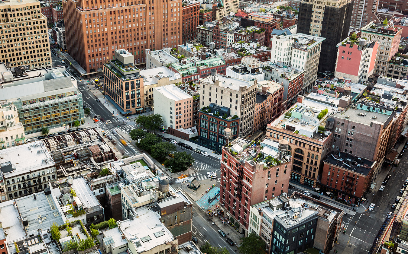 Aerial view of a city block with mid-rise buildings, tree-lined streets, intersections, and cars. Rooftops show various structures and greenery, characteristic of a nyc residential rental neighborhood. The scene has a mix of modern and older architecture.