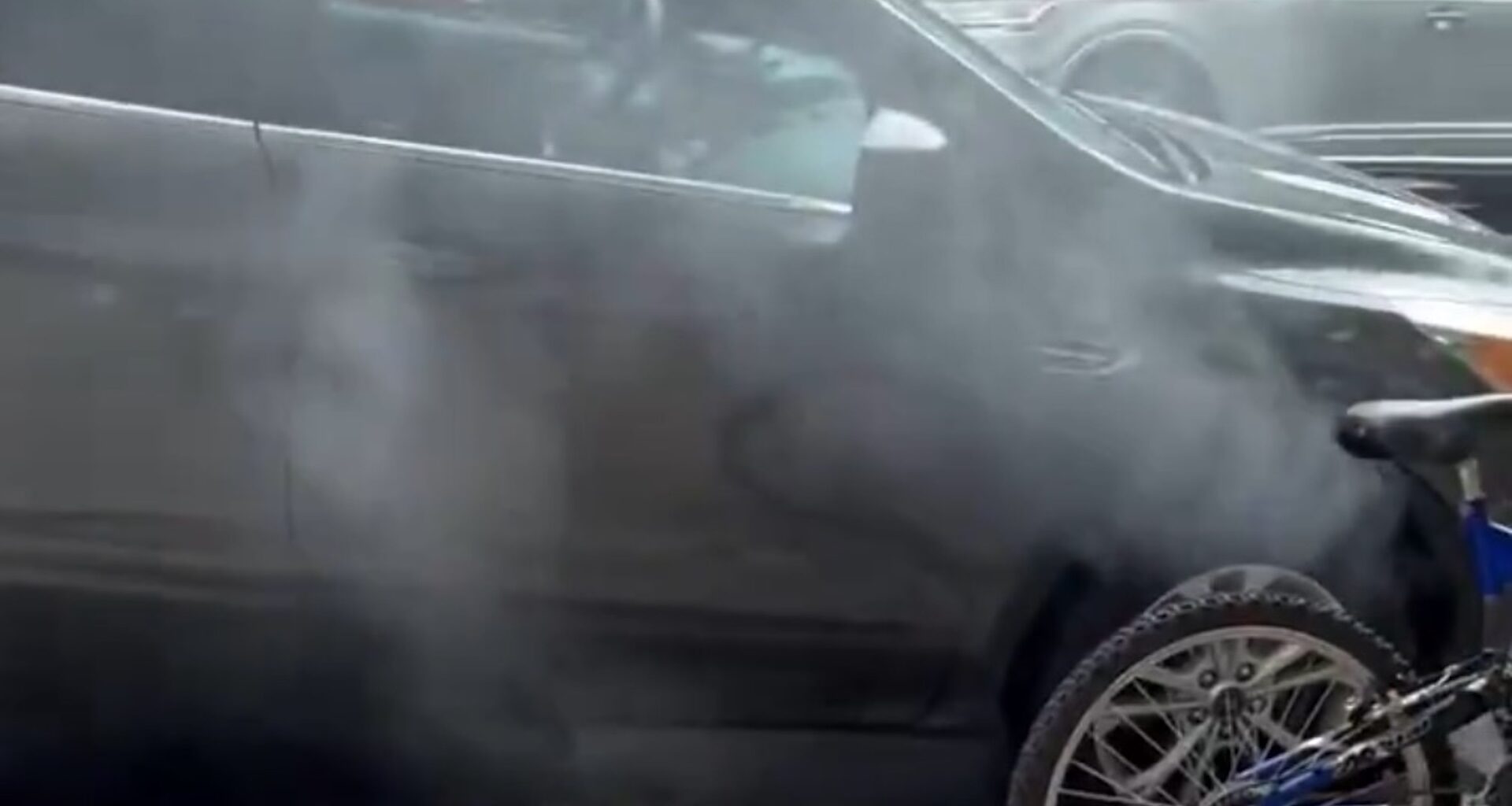Smoke emanates from a parked black vehicle on a street, with a bicycle partially visible in the foreground.