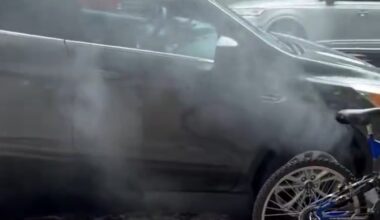 Smoke emanates from a parked black vehicle on a street, with a bicycle partially visible in the foreground.