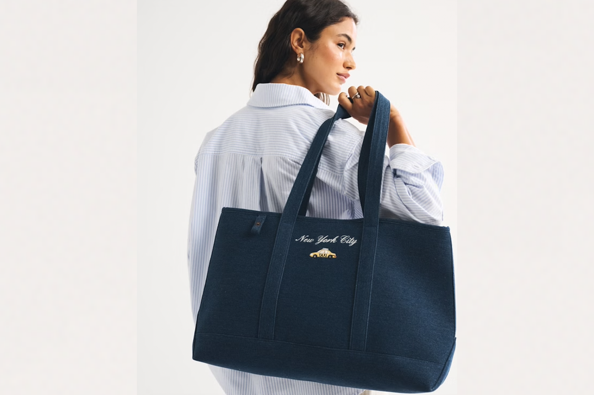 A woman in a blue and white striped shirt carries a denim tote bag with "New York City" and a yellow taxi embroidered on it.