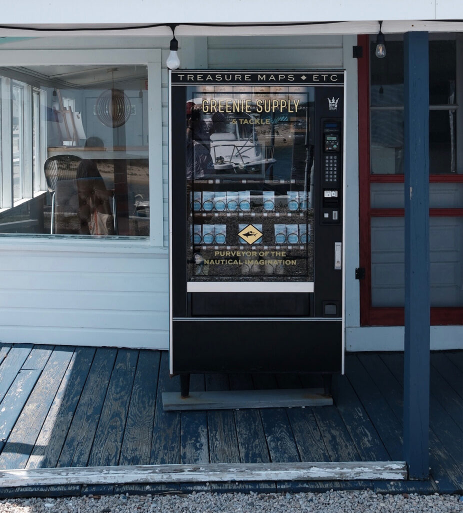 A vending machine labeled “TREASURE MAPS + ETC” stands on a blue wooden porch outside a white building with a red and blue door. The reflection shows a person and nautical decor inside.