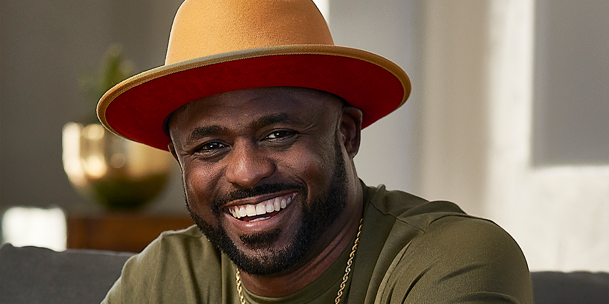 A man with a beard smiles while wearing a tan and orange wide-brimmed hat, a gold chain, and an olive green shirt, seated indoors.