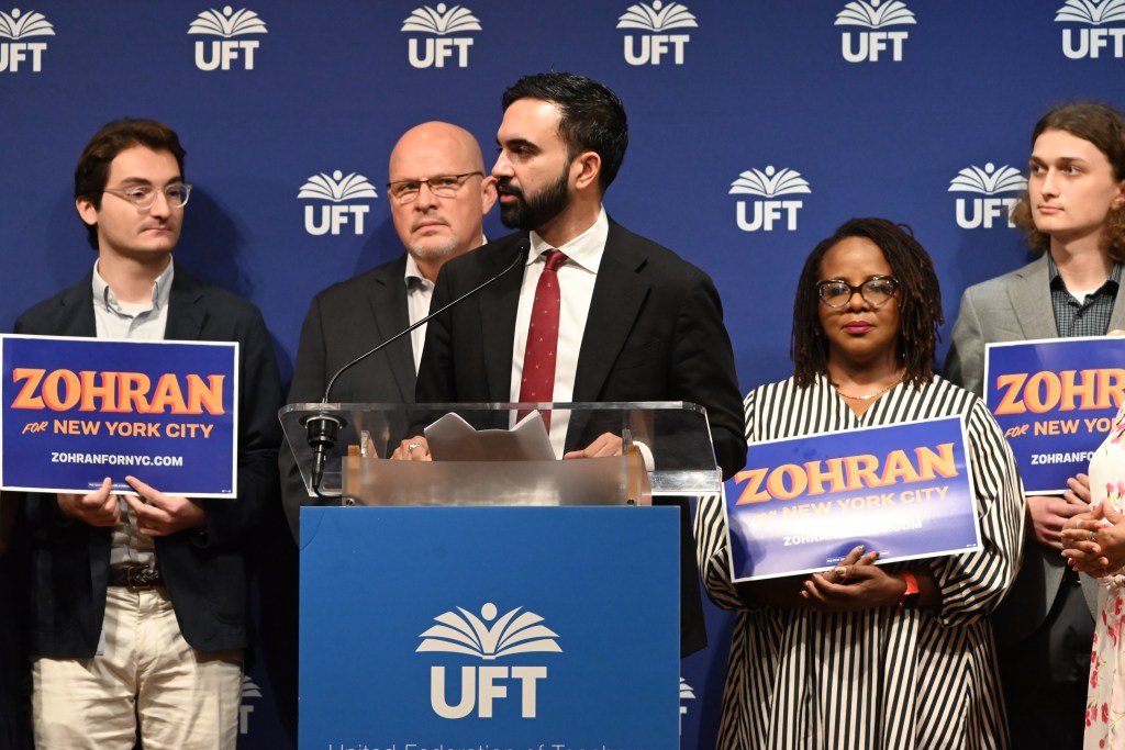 Zohran Mamdani, Democratic nominee for New York City mayor, speaks at a podium with United Federation of Teachers members holding "Zohran for New York City" signs behind him.