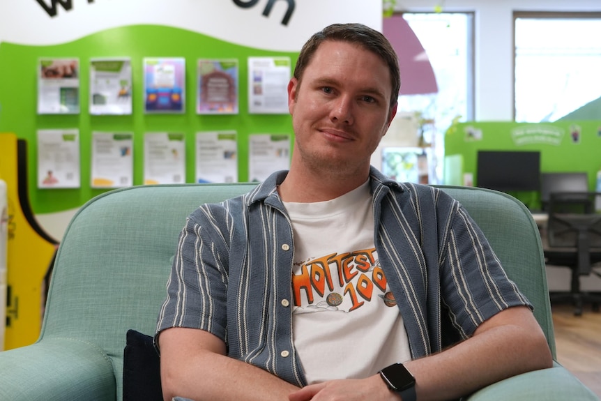 A man (David Myles) sits in a teal chair wearing a white T-shirt and striped button-up shirt over the top.