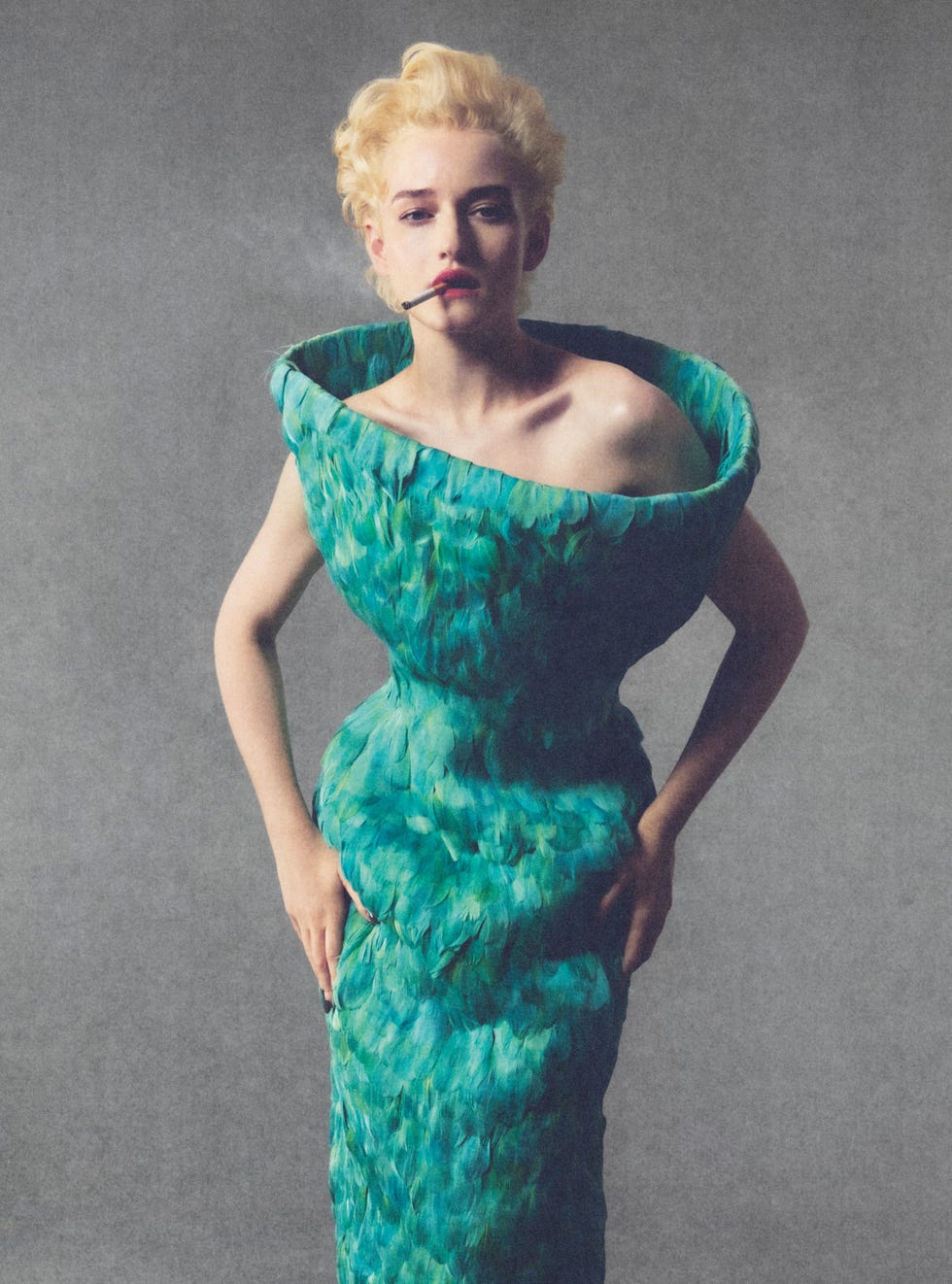 Julia Garner in a textured green dress, posing with a cigarette against a grey background.