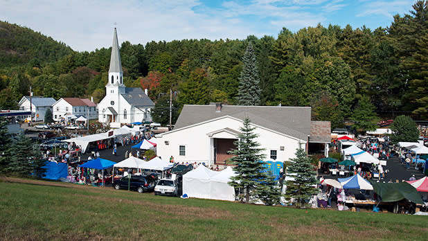 The World's Largest Garage Sale in Warrensburg surrounds a white church with a tall steeple, colorful tents and shoppers set against a forested hillside.