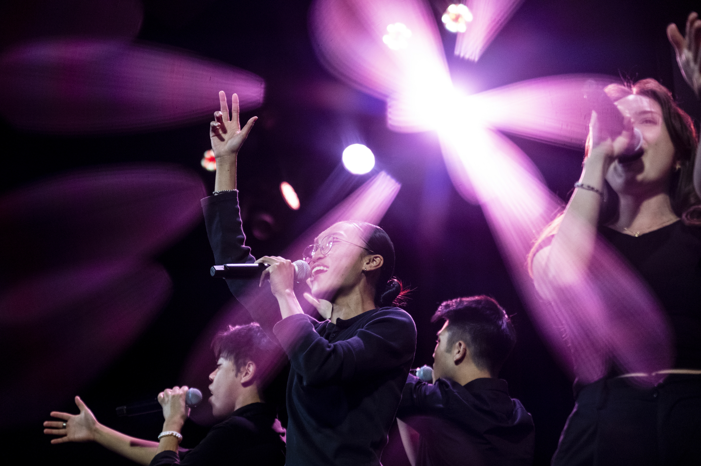 A student performer sings into a microphone and raises a hand onstage as colorful lights illuminate the performance.