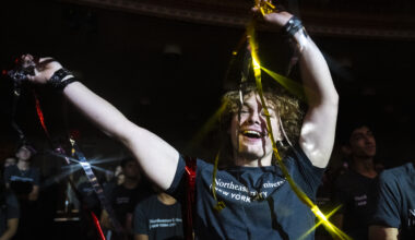 A Northeastern University student in a gray T-shirt raises both arms and smiles while holding metallic gold and red streamers during a lively event at The Town Hall in New York City. Other students stand in the dimly lit auditorium behind them.