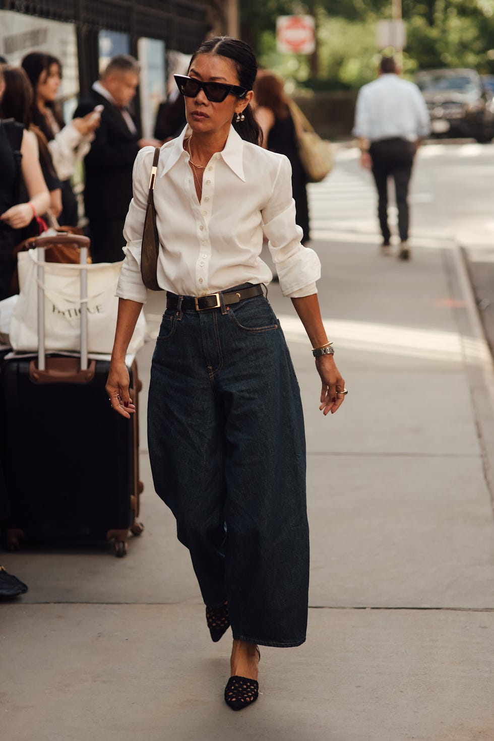 Person walking on the sidewalk wearing a white shirt, wide-leg jeans, and sunglasses, with people and luggage in the background. Person walking on the sidewalk wearing a white shirt, wide-leg jeans, and sunglasses, with people and luggage in the background.