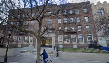 A person rides a bicycle on the sidewalk in front of a large brown brick apartment building with multiple windows and a tree in the foreground.