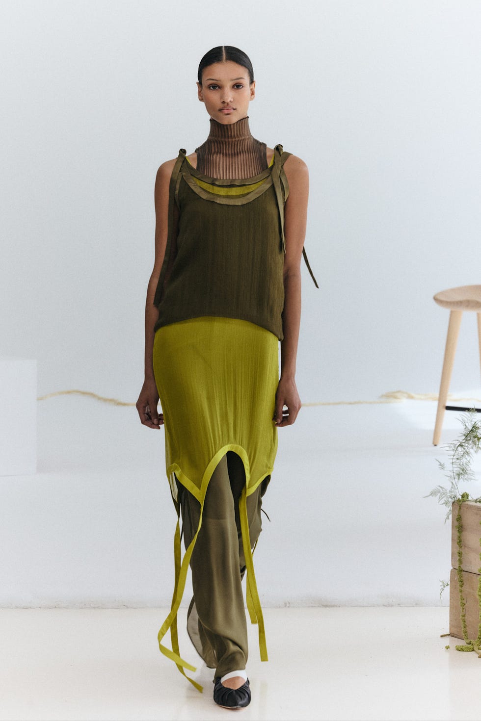 Model wearing a layered outfit in shades of green, with a high neck and unique ribbon details, standing in a minimalist setting.