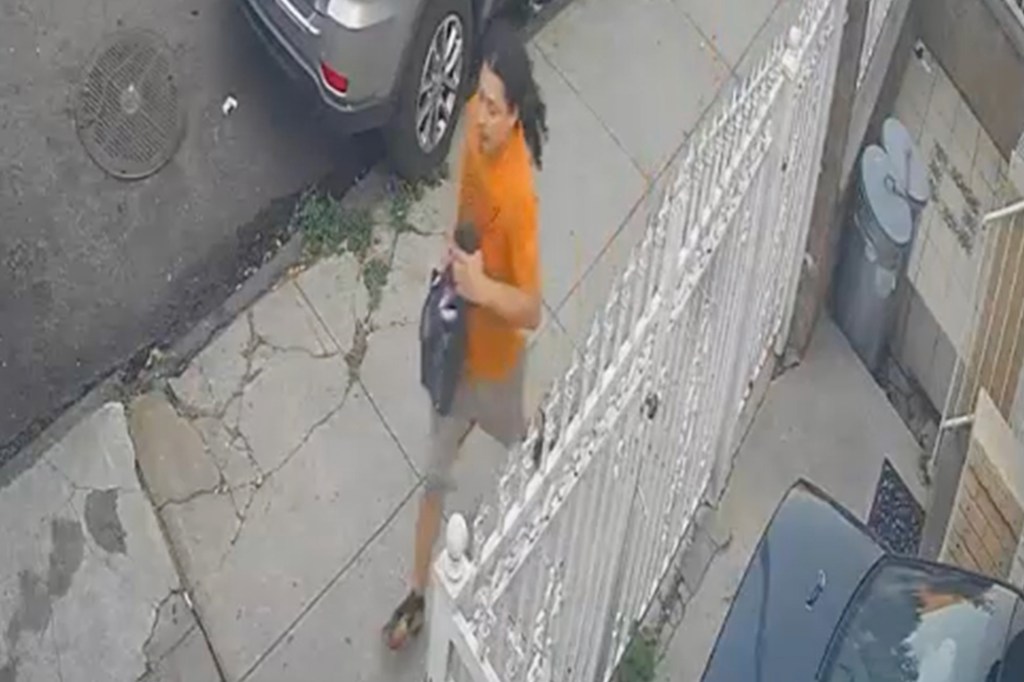 A man in an orange shirt and khaki shorts walks down a sidewalk holding a dark bag.