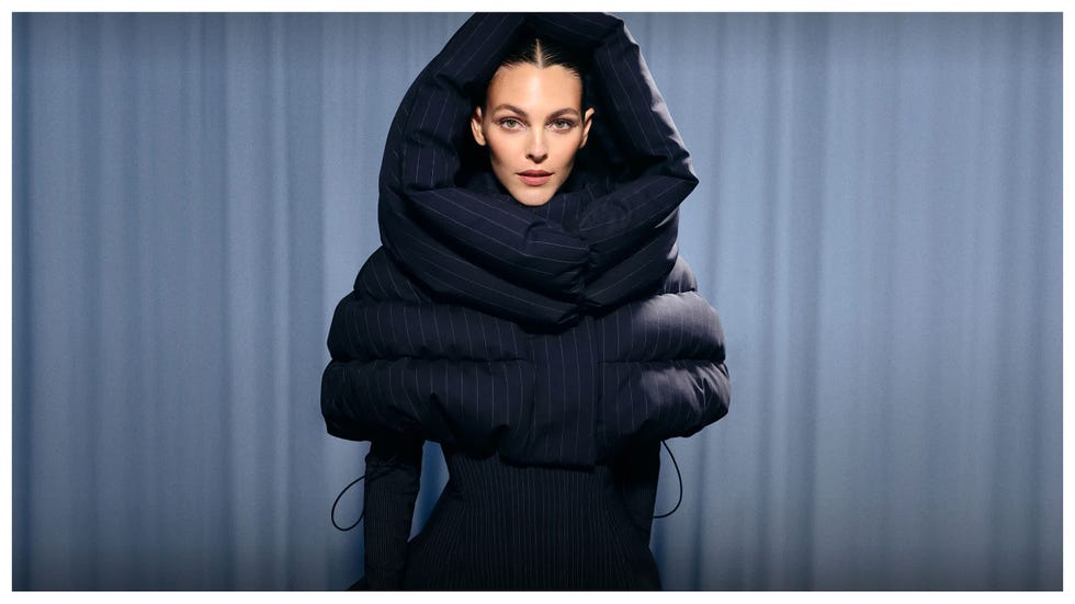 Fashion model wearing a puffer coat with a hood, posing against a soft blue backdrop.