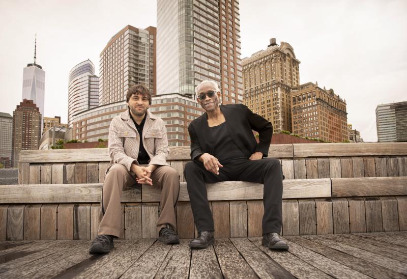 Two men sit in front of the New York City skyline.