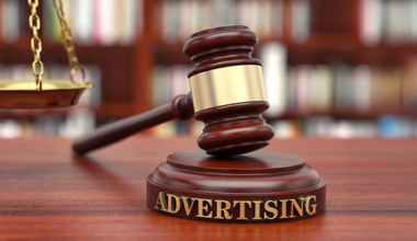 A judges gavel labeled Advertising rests on a wooden desk, with a scale of justice and blurred bookshelves in the background, symbolizing legal issues related to advertising.