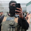 An Immigration and Customs Enforcement officer, wearing tactical face covering and vest, holds up a phone to shoot video.