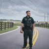 Walton County Sheriff's Office Sgt. Oscar D’Lassalas, wearing his uniform, stands in the middle of a road, on its two yellow parallel dividing lines. A wooden fence is on each side of the road.