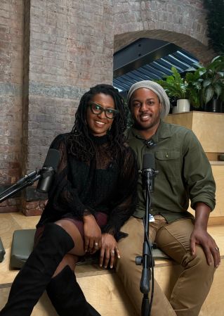 Two people, a woman with dreadlocks and a man with a headscarf, sitting together and smiling at the camera in front of a brick wall.