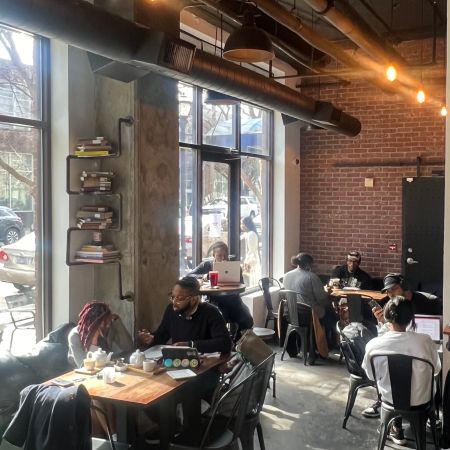 A cozy cafe interior with brick walls, hanging lights, and customers seated at tables, some using laptops.