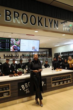 A person standing in a Brooklyn Tea cafe, with the Brooklyn Tea logo and menu visible behind them.