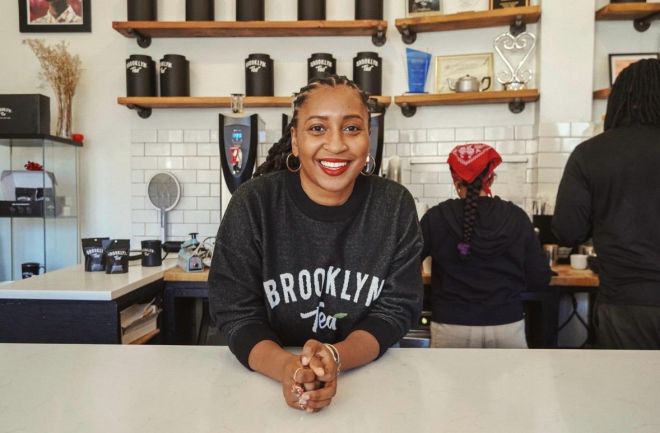 A smiling woman wearing a "Brooklyn Tea" sweatshirt stands behind a counter in a kitchen-like setting, with shelves of tea canisters visible.