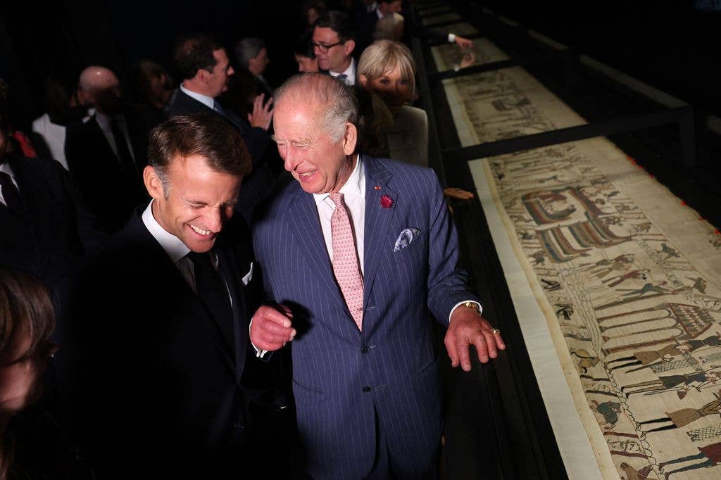 King Charles in a blue suit and pink tie, smiles with another man at an exhibit.