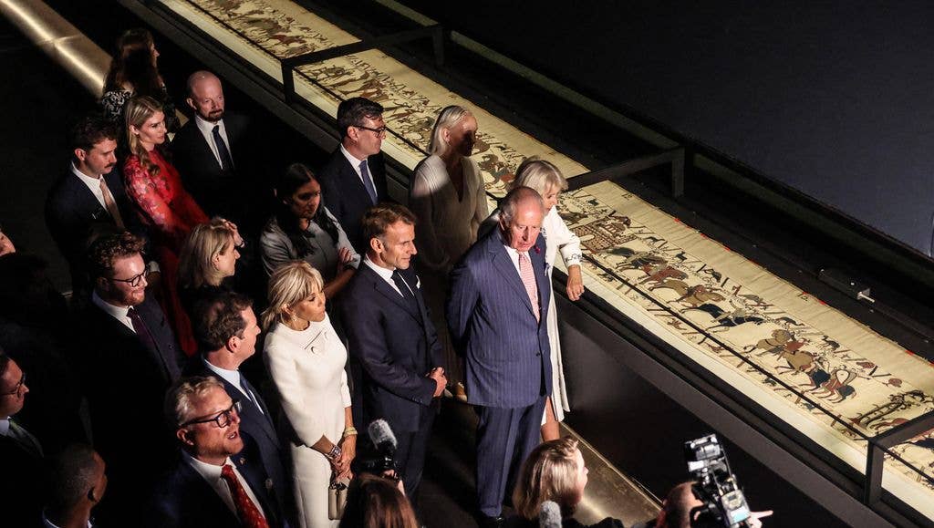 King Charles, in a blue suit, views an exhibit with a group of formally dressed individuals.
