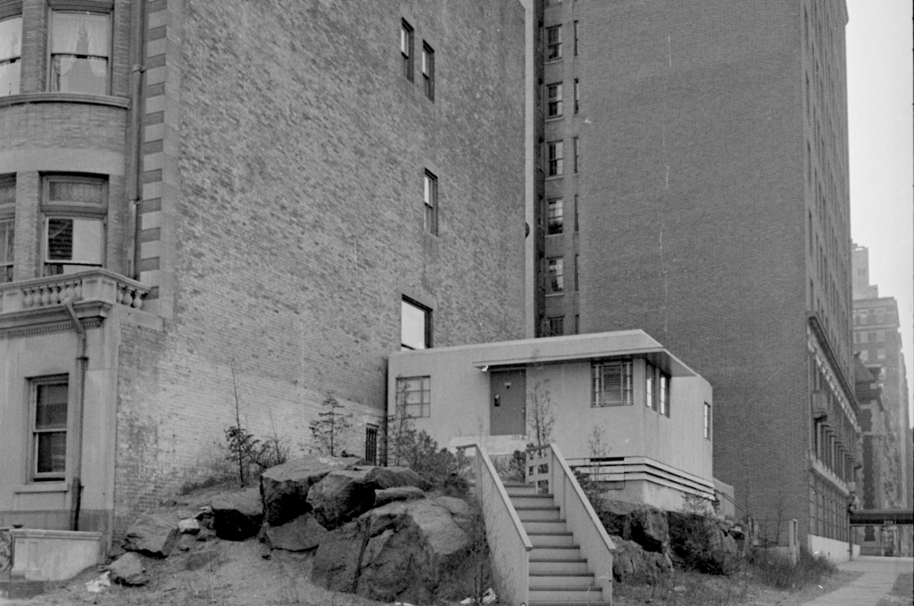 A small, light-colored building situated on a hill of rocks, flanked by larger, brick buildings in a cityscape. The scene is in black and white.