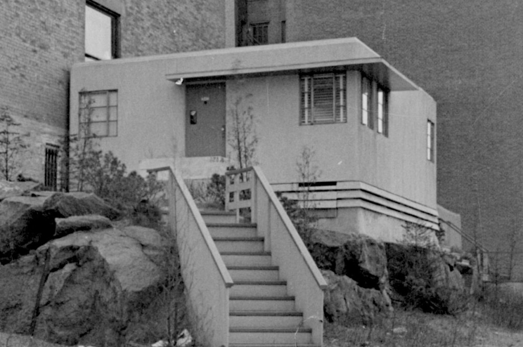 A small, modern house with a flat roof, situated on a rocky hillside, featuring a staircase leading to the entrance and surrounded by a few shrubs.