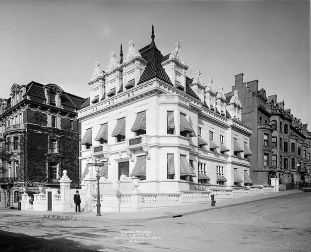 Black and white photograph of a historic building at the corner of a street, featuring elaborate architecture with decorative elements and awnings over the windows.