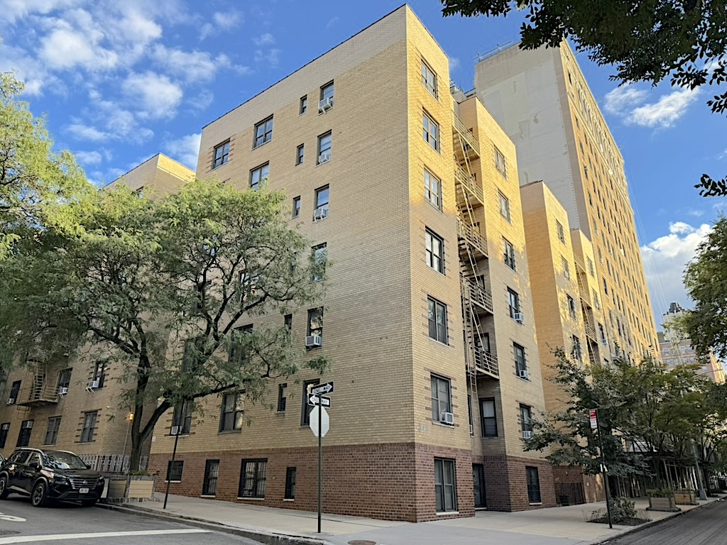 A view of a multi-story residential building with a brick facade, surrounded by trees and a clear blue sky.