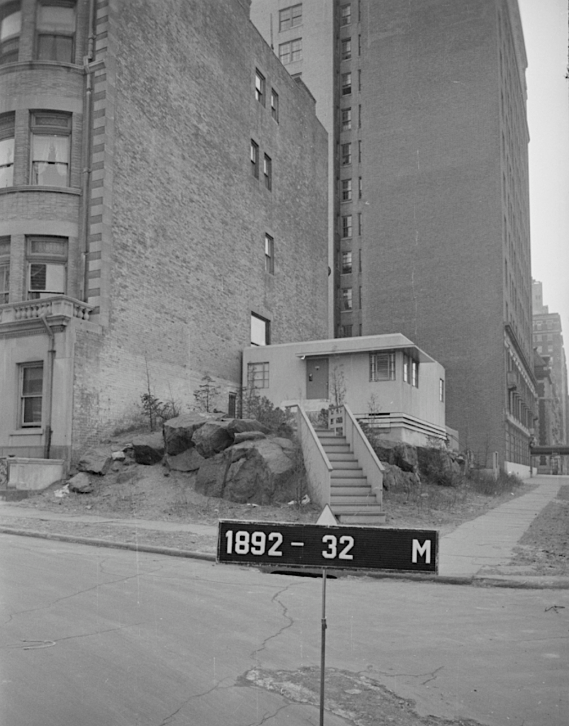 Black and white photograph of a house on a rocky lot beside tall buildings, with a sign displaying '1892 - 32 M' in the foreground.