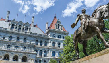 A bronze equestrian statue stands in front of a historic stone building with red-roofed towers, surrounded by trees under a blue sky with clouds.