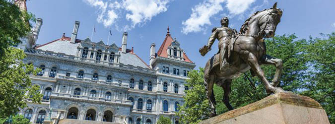 A bronze equestrian statue stands in front of a historic stone building with red-roofed towers, surrounded by trees under a blue sky with clouds.