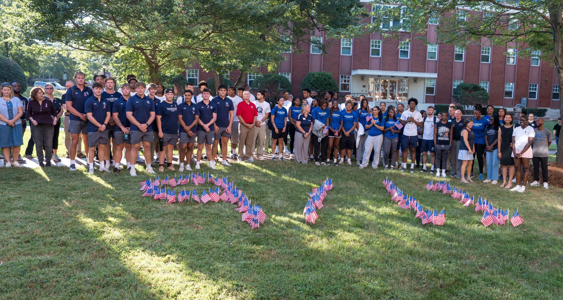 Students, faculty and staff gather on the academic quad lawn behind small American flags in the ground in the shape of 9/11.