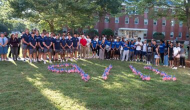 Students, faculty and staff gather on the academic quad lawn behind small American flags in the ground in the shape of 9/11.