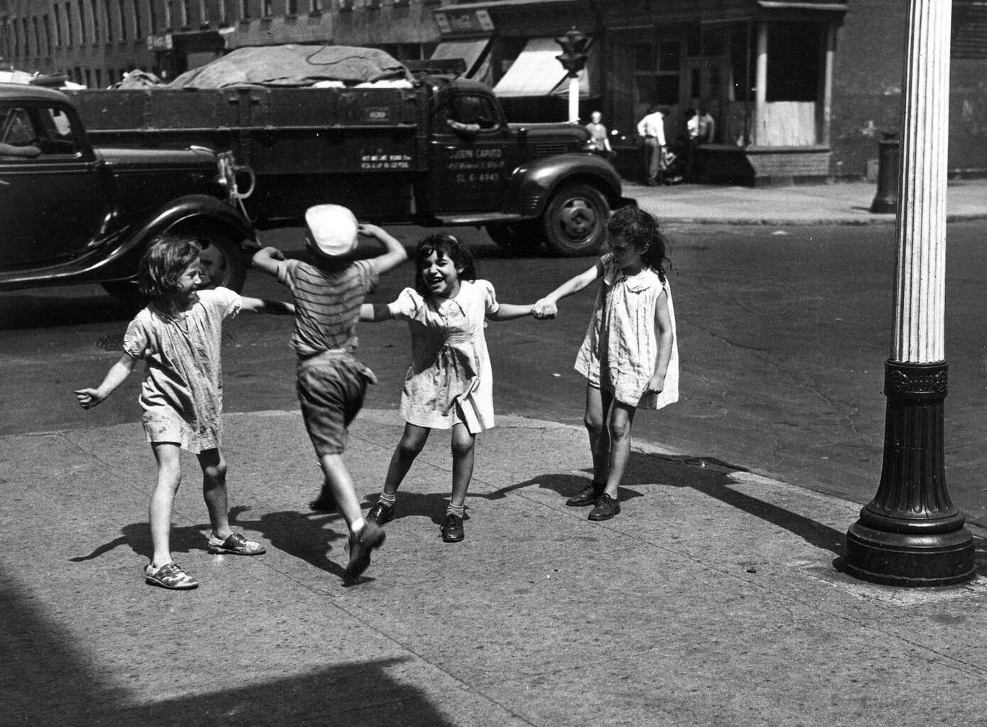 Four children playing together on a city street, two girls holding hands while a boy jumps in the center, with vintage cars and buildings in the background.
