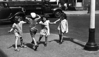 Four children playing together on a city street, two girls holding hands while a boy jumps in the center, with vintage cars and buildings in the background.