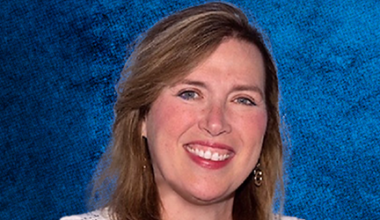 A woman with light brown hair and earrings smiles in front of a textured blue background.