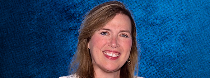 A woman with light brown hair and earrings smiles in front of a textured blue background.