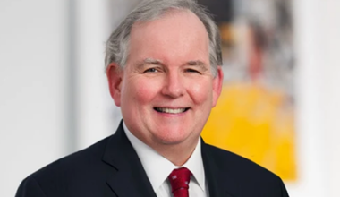 Smiling man with gray hair in a dark suit, white shirt, and red tie, standing in front of a blurred background with white and yellow tones.