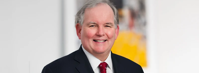Smiling man with gray hair in a dark suit, white shirt, and red tie, standing in front of a blurred background with white and yellow tones.