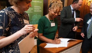 A woman in a green dress signs a document at a podium labeled American Irish Historical Society, while three others stand nearby, engaged and holding papers.
