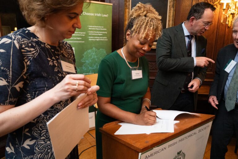 A woman in a green dress signs a document at a podium labeled American Irish Historical Society, while three others stand nearby, engaged and holding papers.
