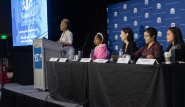 A speaker stands at a podium addressing a panel of four seated women at a conference. A blue screen and banner displaying Partnership Conference are visible in the background. Nameplates and microphones are on the table.