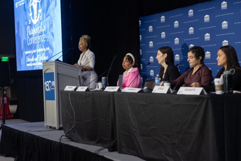 A speaker stands at a podium addressing a panel of four seated women at a conference. A blue screen and banner displaying Partnership Conference are visible in the background. Nameplates and microphones are on the table.