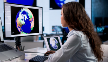 A woman in an office studies climate data maps of Earth on two large monitors, analyzing colorful weather patterns and geographic information, with a laptop and another screen nearby.